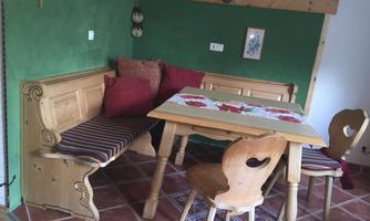 A cozy dining area with a wooden bench, a table, and chairs. The wall is painted green, and there are decorative cushions and pictures.