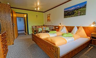A cozy bedroom with a large double bed and wooden details. In the background, there is a dining table and a wall picture of a mountain landscape.