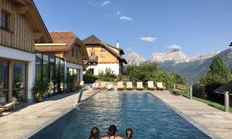 A beautiful pool overlooking the mountains and traditional wooden houses. Three people are relaxing by the edge of the pool under a clear sky.