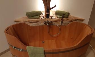 A rustic wooden bathtub stands in a modern room. Above the tub, two green towels and a bottle of wine are placed on an elegant shelf.