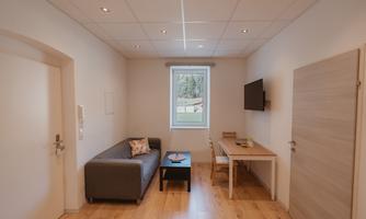 A bright room with a gray sofa, a table, and a television. Large windows provide plenty of natural light.