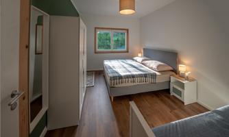 A modern bedroom with a double bed and a single bed. The room has light walls, a wooden floor, and large windows.