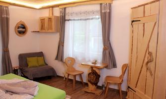 A cozy room with a bed, a gray sofa, and a small wooden table. The windows are decorated with curtains and there is a wooden chest as an accent.