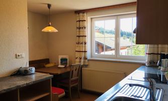 A bright kitchen with a dining table and windows that offer a view of a green landscape. The decor is simple and cozy.