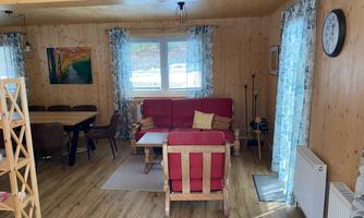 A cozy living room with wooden walls and a red sofa. The room is well-lit and has large windows with curtains.