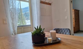 A cozy room with a wooden table, on which sits a plant arrangement and candles. The view from the window shows a green landscape.