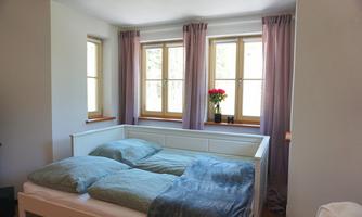 A bright bedroom with a white bed and colorful cushions. Large windows with purple curtains and a vase with flowers create a fresh atmosphere.