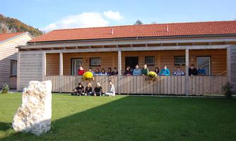 A group of people is standing in front of a wooden house with red roofs. In the foreground, there is a large stone on the green meadow.