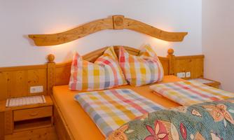 A cozy bedroom with a wooden bed and colorful pillows. The walls are bright, and there is a small nightstand next to the bed.