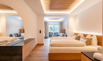 A modern hotel room with a large double bed and soft lighting. In the background, there is a cozy seating area and a balcony with a view.