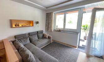 A modern living room with a gray sofa and a carpet. Large windows let in plenty of light and show a view of the outdoor area.