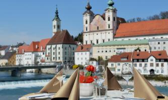 A beautifully decorated table with glasses and fresh strawberries stands by the river. In the background, historic buildings and a picturesque landscape can be seen.