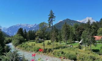 A picturesque landscape with mountains in the background and a clear blue sky. A river flows through the green meadow, surrounded by trees and colorful flowers.