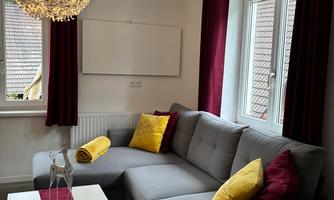 A cozy living room with a gray corner sofa and colorful cushions. The windows are adorned with red curtains, and a striking lamp provides atmospheric lighting.