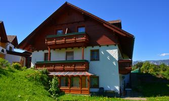 A two-story house with a beautiful balcony and a spacious window front. It is situated in a green environment under a blue sky.