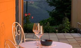 A cozy table with two chairs on a terrace, surrounded by mountains. Two wine glasses and a bowl are on the table in the warm light of the sunset.
