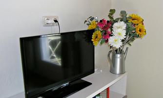A television is on a white shelf. Next to it is a vase with colorful artificial flowers.