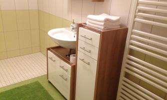 A modern bathroom with a sink and a wooden cabinet. The walls are light tiled and there is a green carpet on the floor.