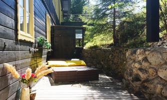 A cozy outdoor area with wooden decking and a rattan furniture set. The surroundings are green with trees and an inviting entrance.