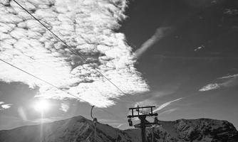 A tall mountain with snow-covered slopes and a cable car in the foreground. The sky is filled with clouds and beams of sunlight.
