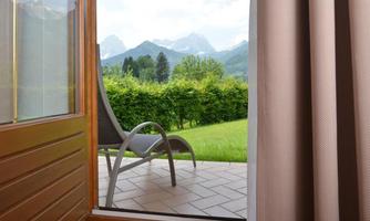 View from a window onto a green meadow with a chair and mountains in the background. The sky is cloudy but friendly.