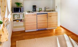 A small, modern kitchen with wooden shelves and a sink. The floor is covered with light parquet, and a rug lies next to the kitchenette.