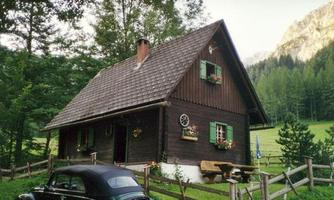 A traditional wooden house in nature, surrounded by trees and meadows. An old wagon stands in front of it, while the mountains are visible in the background.