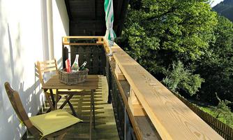 A cozy balcony with a wooden railing and two chairs. On the table, there is a bottle and a basket, while the green nature is visible in the background.