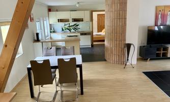 A bright and modern apartment with a dining area and an open kitchen. The living room is equipped with a television and a cozy bedroom in the background.