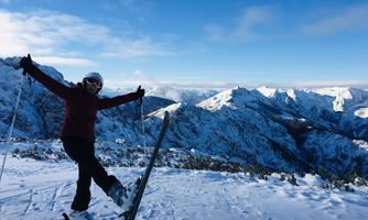 A female skier stands on a snow-covered mountain peak and is happy. In the background, there are snow-covered mountains and a blue sky visible.