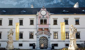 A historical building with a decorative facade and large windows. Two statues stand on either side of the entrance, while yellow banners hang in the foreground.