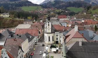 A picturesque town landscape with historic buildings and a church in the center. In the background, we see green mountains and a calm, idyllic setting.