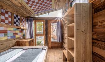 A cozy bedroom with wooden-paneled walls and colorful patterns. The large windows let in plenty of daylight and offer a view of a garden.