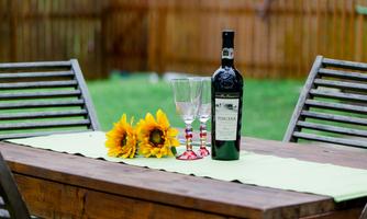 A wooden table outdoors with a bottle of red wine, two glasses, and a group of sunflowers. The table is decorated with a green table runner, and a wooden wall is visible in the background.