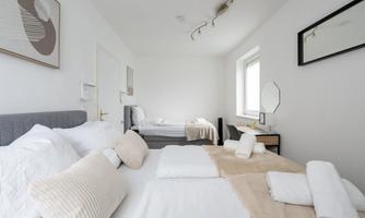 A bright, modern bedroom with a large bed and a single bed. The decor is simple and inviting, with neutral colors and decorative cushions.
