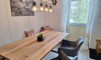A cozy dining area with a large wooden table and modern chairs. Large windows let in plenty of light, and there is a plant as decoration.