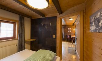 A cozy bedroom with wooden paneling and a window. In the background, a dining area can be seen.