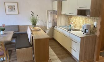 A modern kitchen with wooden shelves and a cozy seating area. The room is bright and inviting, featuring a refrigerator and a small table.