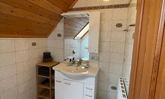 A bathroom with slanted wood paneling and tiles. There is a sink, a mirror, and a small wooden shelf.