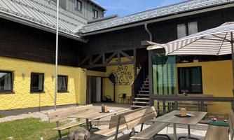 A cozy courtyard with wooden terraces and a sun umbrella. The walls are painted in yellow and dark brown, and there is a staircase leading to an upper area.