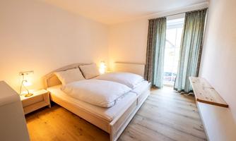 A bright bedroom with a large bed and two pillows. Large windows let in a lot of light, and there is a cozy wooden shelf.