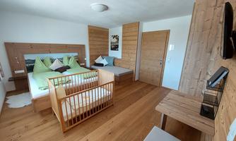A cozy bedroom with wooden paneling and a double bed. In the corner stands a baby crib, and there is a small desk.