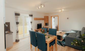 A modern living room with a dining table and blue chairs. The large window front lets in a lot of light and offers access to a balcony.