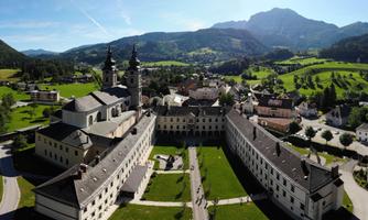 A picturesque monastery complex surrounded by green meadows and mountains. The sky is clear and the architecture is impressive.