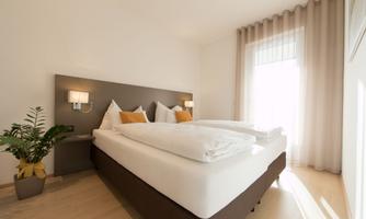 A bright bedroom with a large double bed and two pillows. The room has windows with curtains and a small plant in the corner.