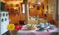 A cozy kitchen with wooden furniture and a set table. On the table are flowers, dishes, and bread.
