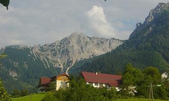 A picturesque mountain landscape with majestic peaks and a tranquil farmhouse in the foreground. The nature is lush and green, under a slightly cloudy sky.