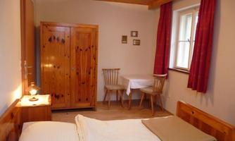 A cozy bedroom with a wooden wardrobe, two chairs, and a table. The walls are light, and the windows have red curtains.