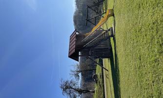 A playground with a slide and a climbing frame is situated on a green lawn. In the background, trees and gentle hills are visible.