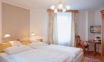 A cozy bedroom with two single beds and a bright window front. The room is stylishly furnished with a lamp and a table chair.